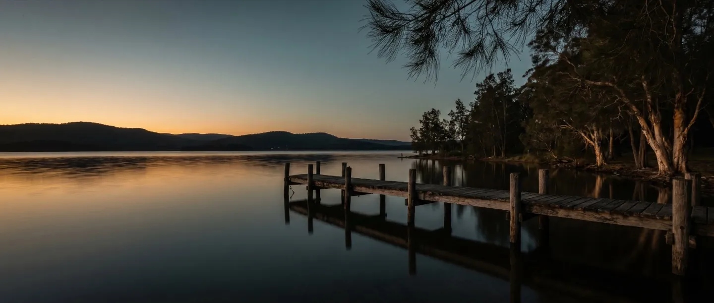 Still lake water at dusk from the bush shoreline, an old timber jetty reaching into the water